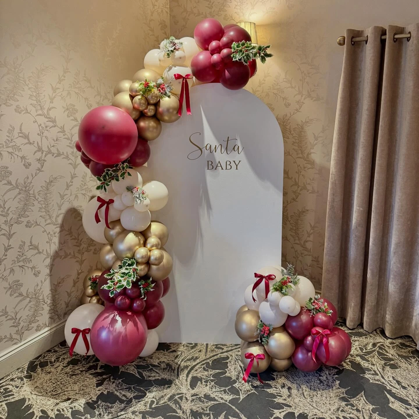 Red and gold Santa Baby balloon arch backdrop with greenery and neutral curtains