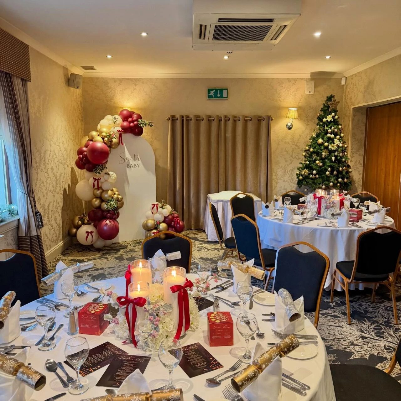 Elegant Santa Baby event room with round tables, candles, a balloon arch, and a Christmas tree