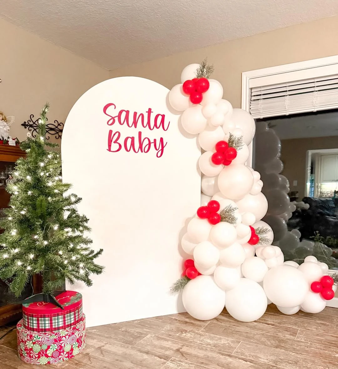 Minimalist Santa Baby balloon arch with white balloons, red details, pine greenery, and gift boxes