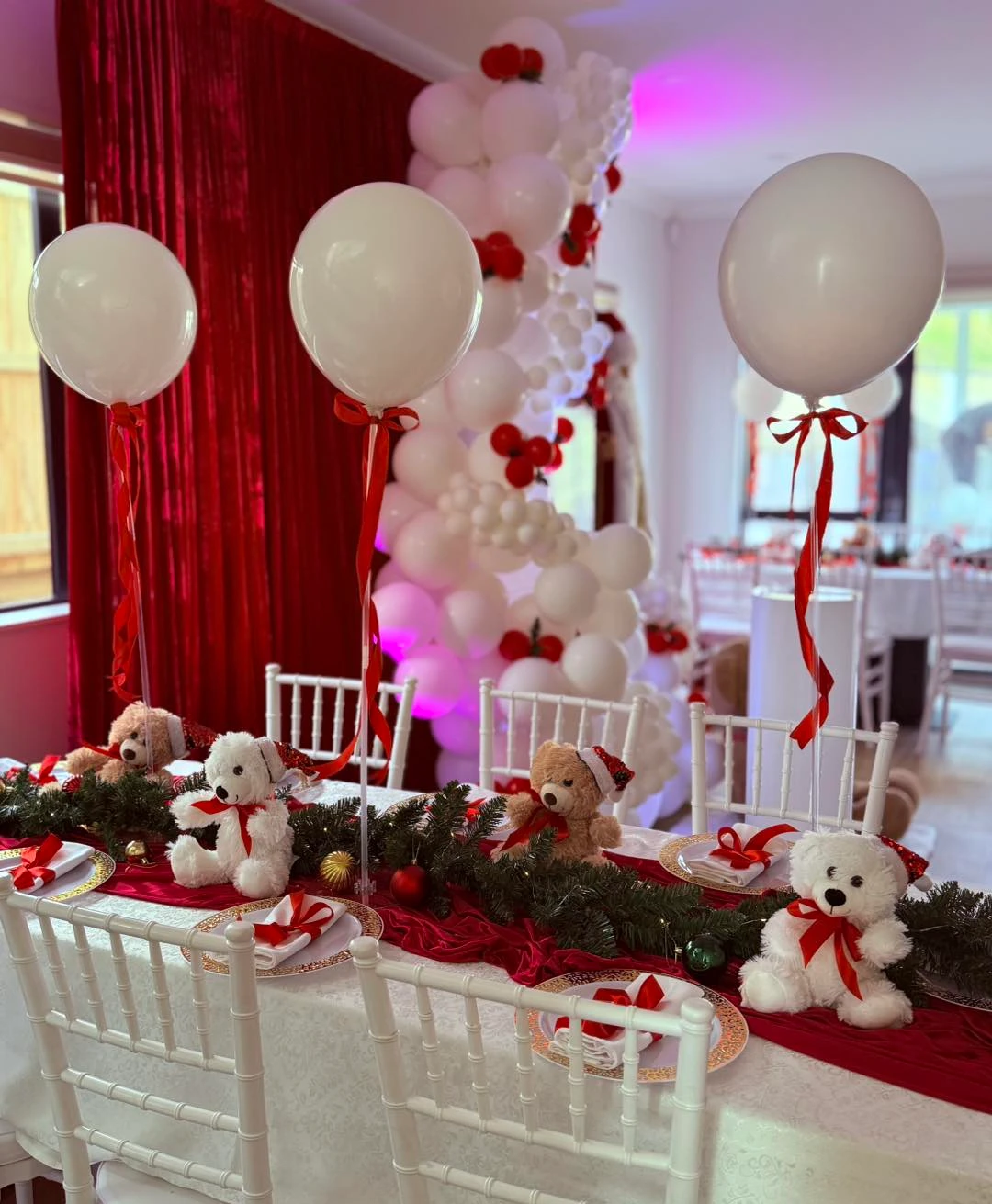 Plush bear dining display with teddy bears, white balloons, red bows, pine branches, and gold ornaments